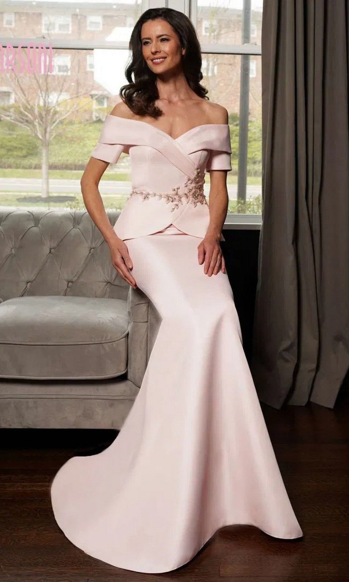 A woman wearing the Marsoni MV1144 long formal dress in blush, featuring an off-the-shoulder neckline, beaded peplum waistline, fitted bodice, and flared skirt, sits on a couch in a room with large windows.
